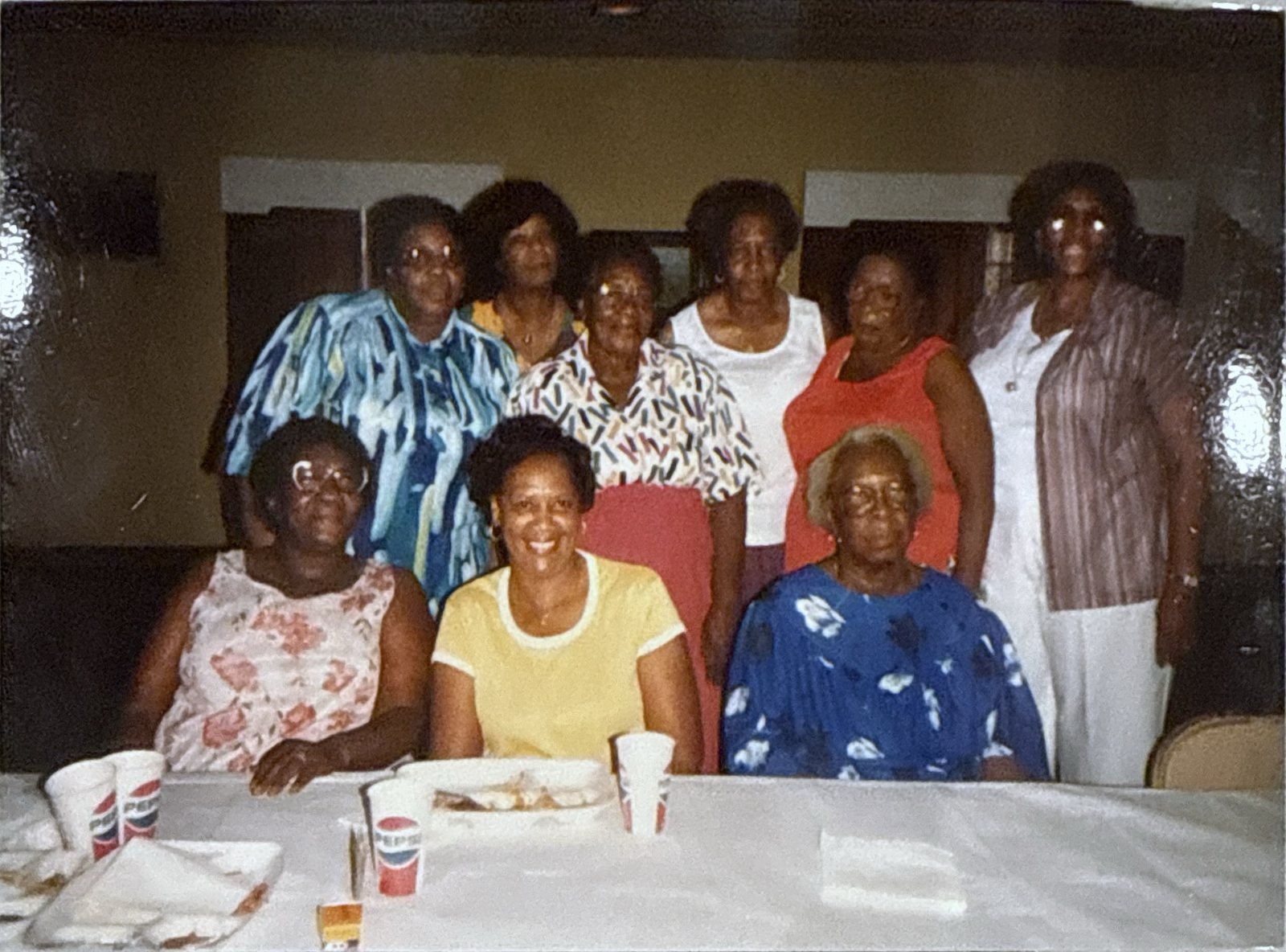 Older family photograph of ten women posed together, three seated at a table set with paper cups