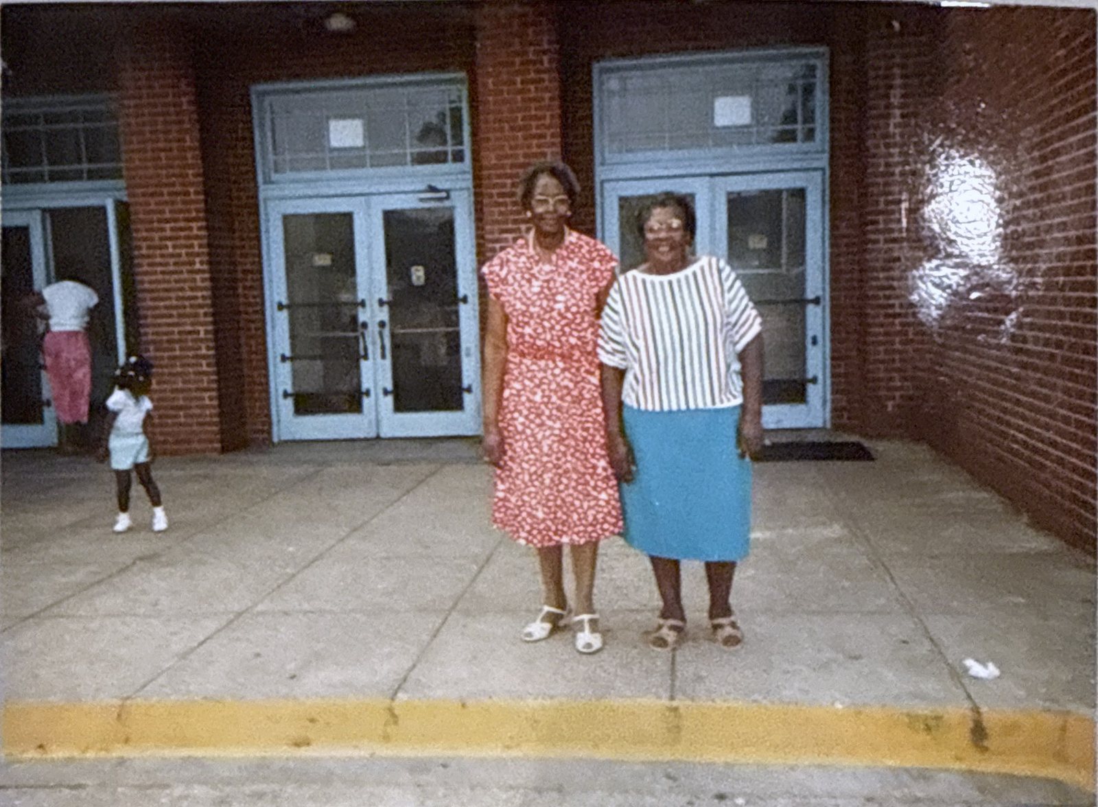 Older photograph of two women standing together outside a brick building, a small child playing nearby